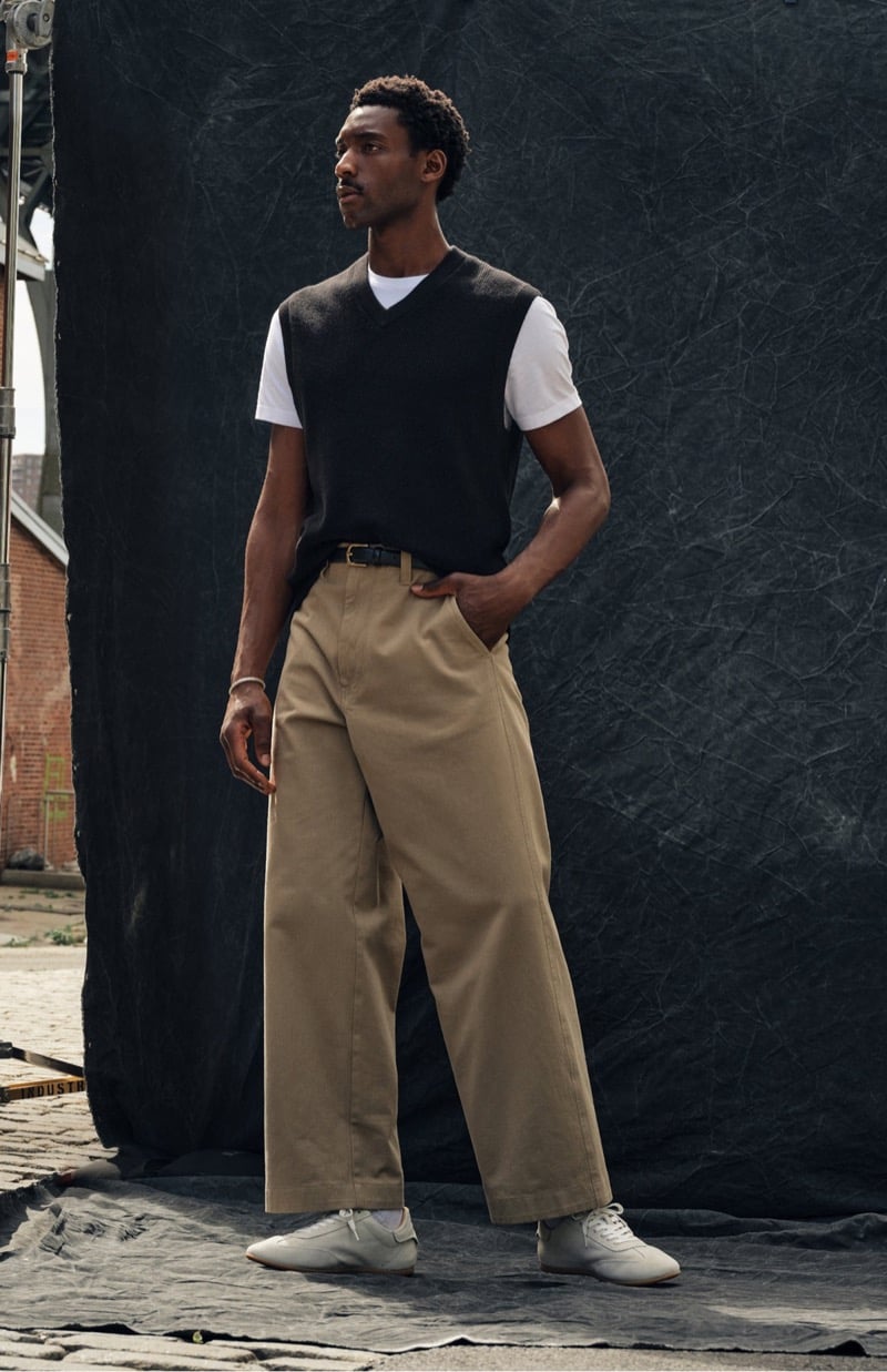 Halid Nuhu stands with a hand in his pocket in extra-wide khaki chinos with a V-neck sweater vest, a tee, and low-top sneakers, for Todd Snyder's Essential Pieces