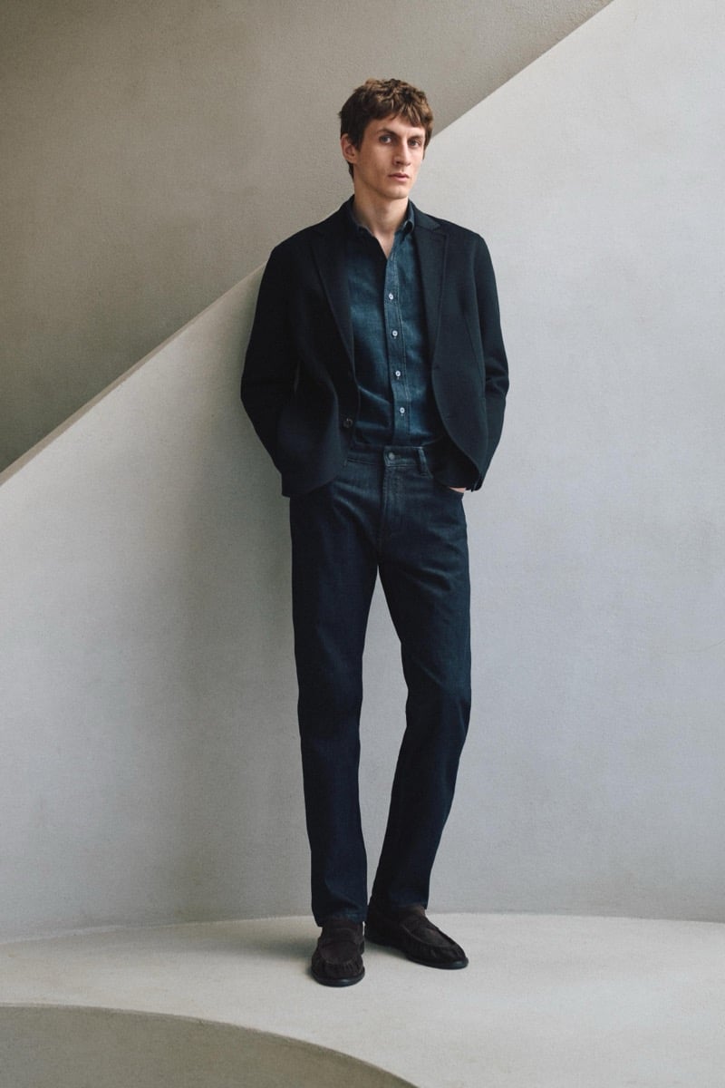 Henry Kitcher stands against a curved wall in an unstructured blazer over a denim shirt with dark jeans and suede loafers, for Massimo Dutti's The Loft