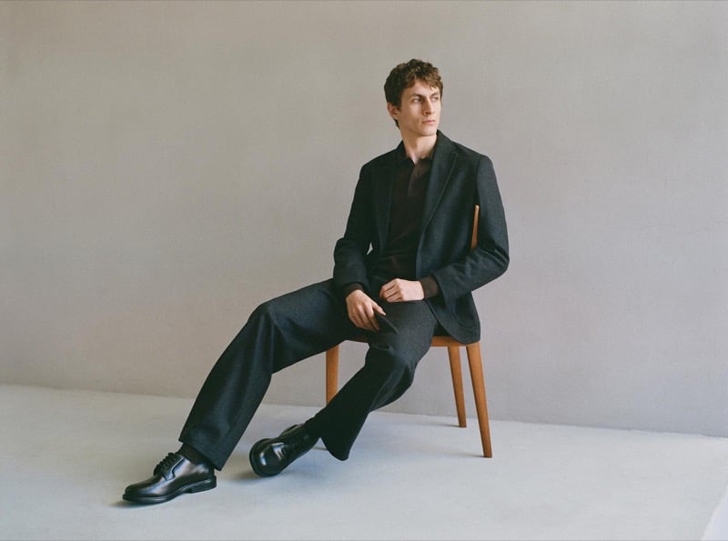 Henry Kitcher sits on a wooden chair in a textured blazer and knit polo with wide trousers and black derby shoes against a gray backdrop, for Massimo Dutti's The Loft