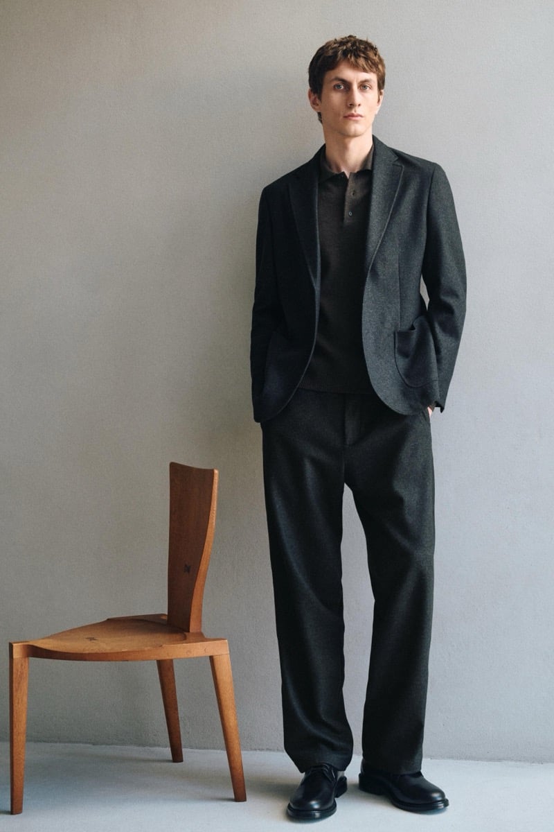 Henry Kitcher stands with a hand in his pocket in a textured blazer over a knit polo with wide trousers and black derby shoes beside a wooden chair, for Massimo Dutti's The Loft
