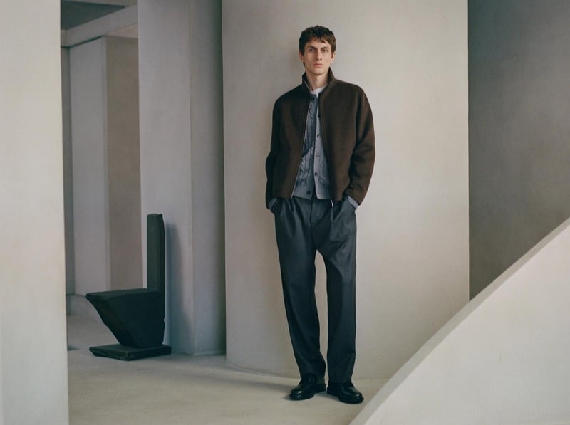 Henry Kitcher stands with his hands in his pockets against a curved plaster wall in a brown zip jacket over a gray cable-knit cardigan with pleated trousers and black lace-up shoes, for Massimo Dutti's The Loft