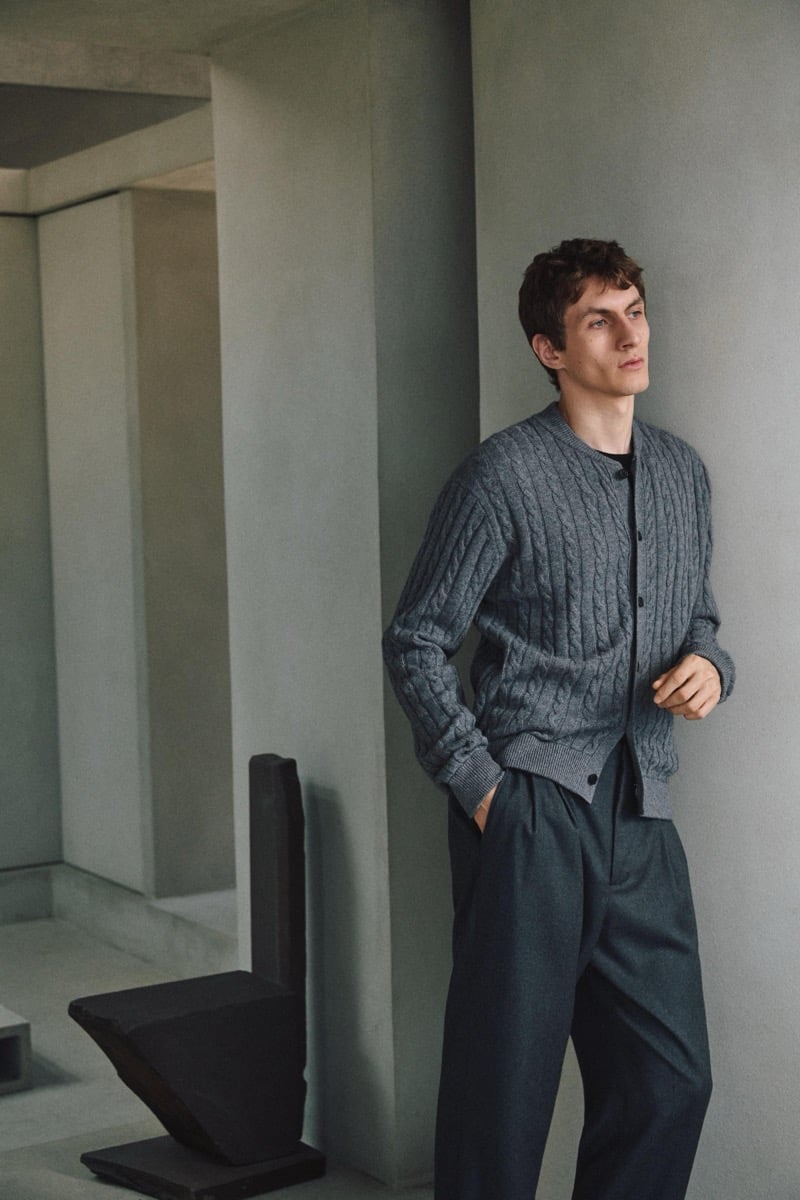 Henry Kitcher leans against a plaster wall in a gray cable-knit cardigan with pleated trousers, a sculptural chair behind him, for Massimo Dutti's The Loft