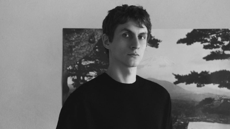Black-and-white portrait of Henry Kitcher in a crewneck sweater in front of a framed landscape photograph of a mountain and pine trees, for Massimo Dutti's The Loft