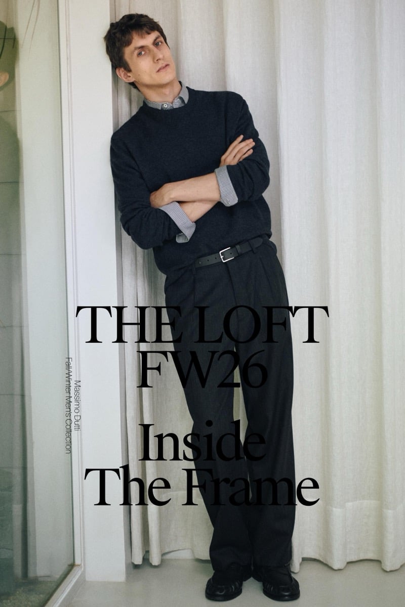 Henry Kitcher leans against a wall with his arms crossed in a crewneck sweater over a checked shirt with wide pleated trousers and black shoes, with The Loft FW26 title text over the image, for Massimo Dutti's The Loft