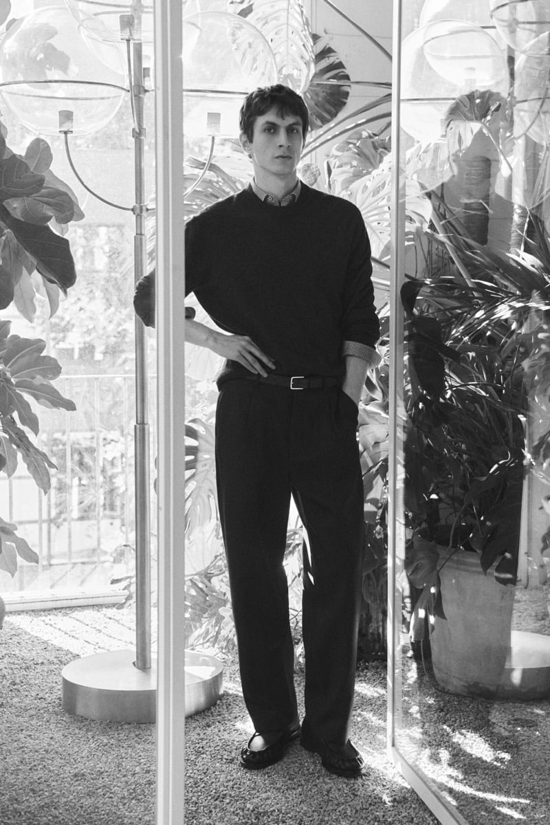 Black-and-white image of Henry Kitcher standing among plants behind glass panels in a crewneck sweater over a checked shirt with pleated trousers and loafers, for Massimo Dutti's The Loft