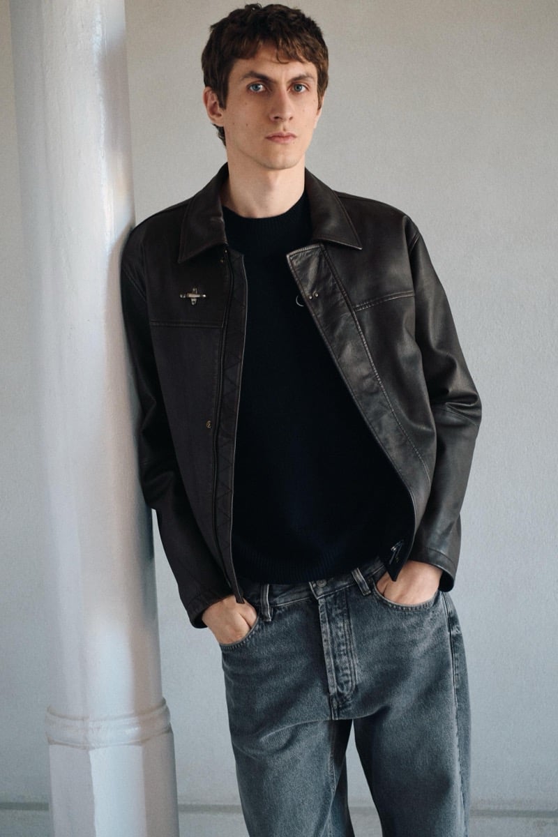 Henry Kitcher leans against a painted column in an open leather jacket over a dark crewneck sweater with washed jeans, for Massimo Dutti's The Loft