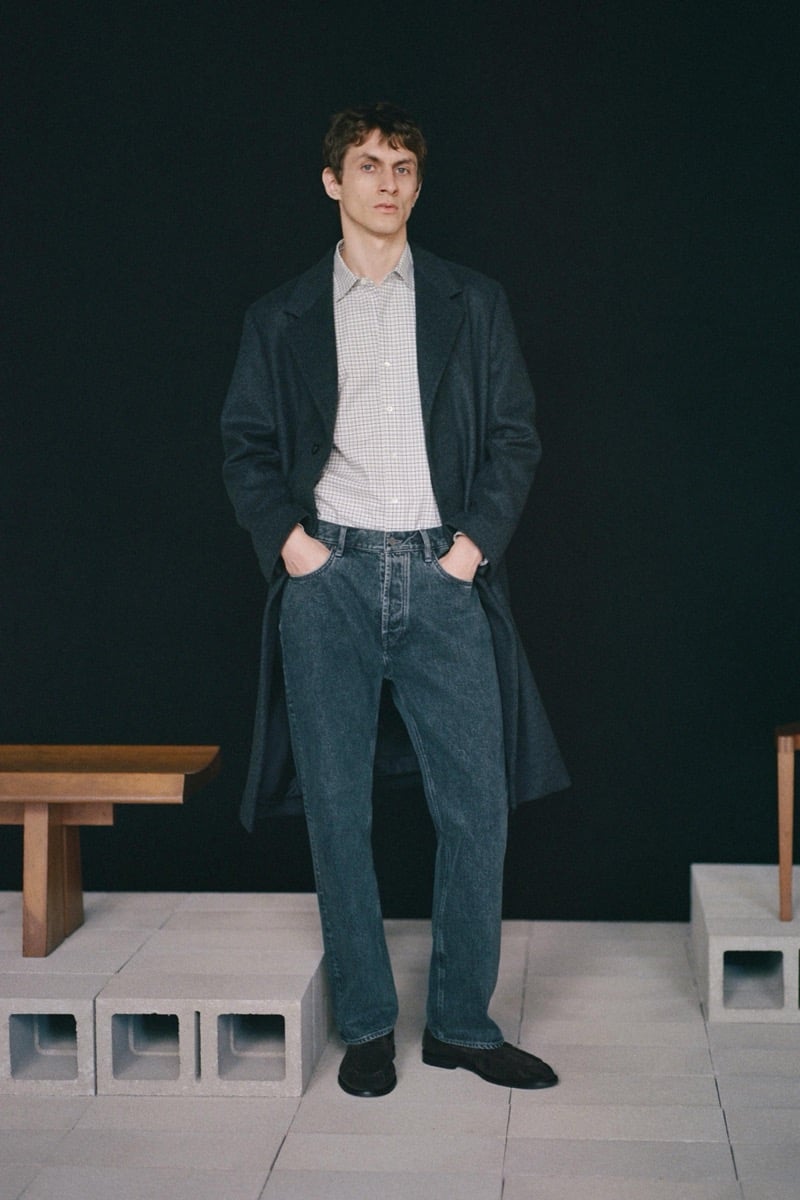 Henry Kitcher stands with his hands in his pockets in a long overcoat over a checked shirt with washed jeans and suede loafers against a dark backdrop, for Massimo Dutti's The Loft