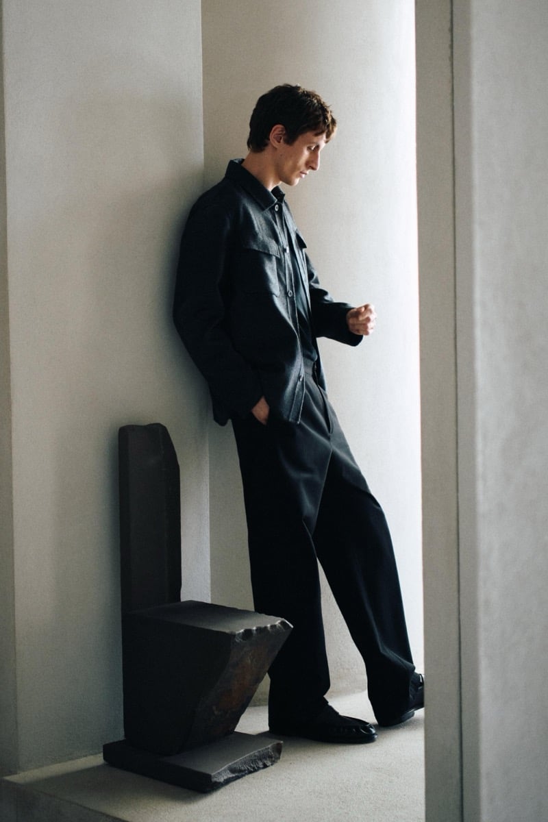 Henry Kitcher leans against a wall beside a sculptural stone chair in a chest-pocket overshirt and wide trousers with loafers, for Massimo Dutti's The Loft