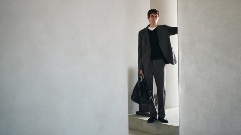 Henry Kitcher stands in a plaster doorway in an oversized blazer over a V-neck sweater and tee with pleated trousers, a black bag in hand, for Massimo Dutti's The Loft