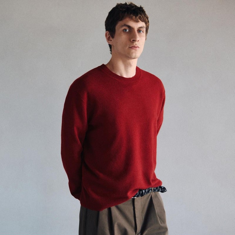 Henry Kitcher wears a red crewneck sweater with pleated trousers and a woven belt against a plain wall, for Massimo Dutti's The Loft