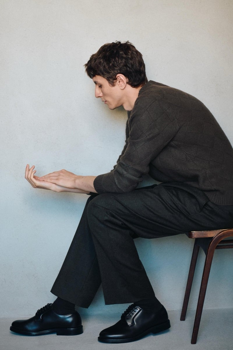 Henry Kitcher sits in profile on a wooden stool in a textured knit sweater and wide trousers with black derby shoes, for Massimo Dutti's The Loft