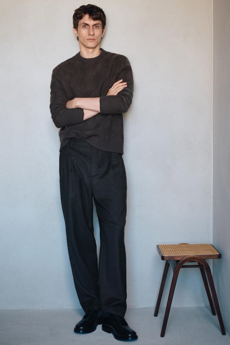 Henry Kitcher stands with his arms crossed in a textured knit sweater and wide pleated trousers with black derby shoes beside a woven-seat stool, for Massimo Dutti's The Loft