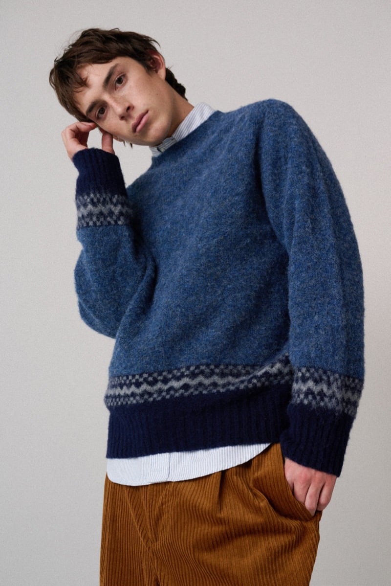 A model in a blue sweater with patterned trim at the cuffs and hem, paired with corduroy trousers, for Howlin's fall-winter 2026 collection