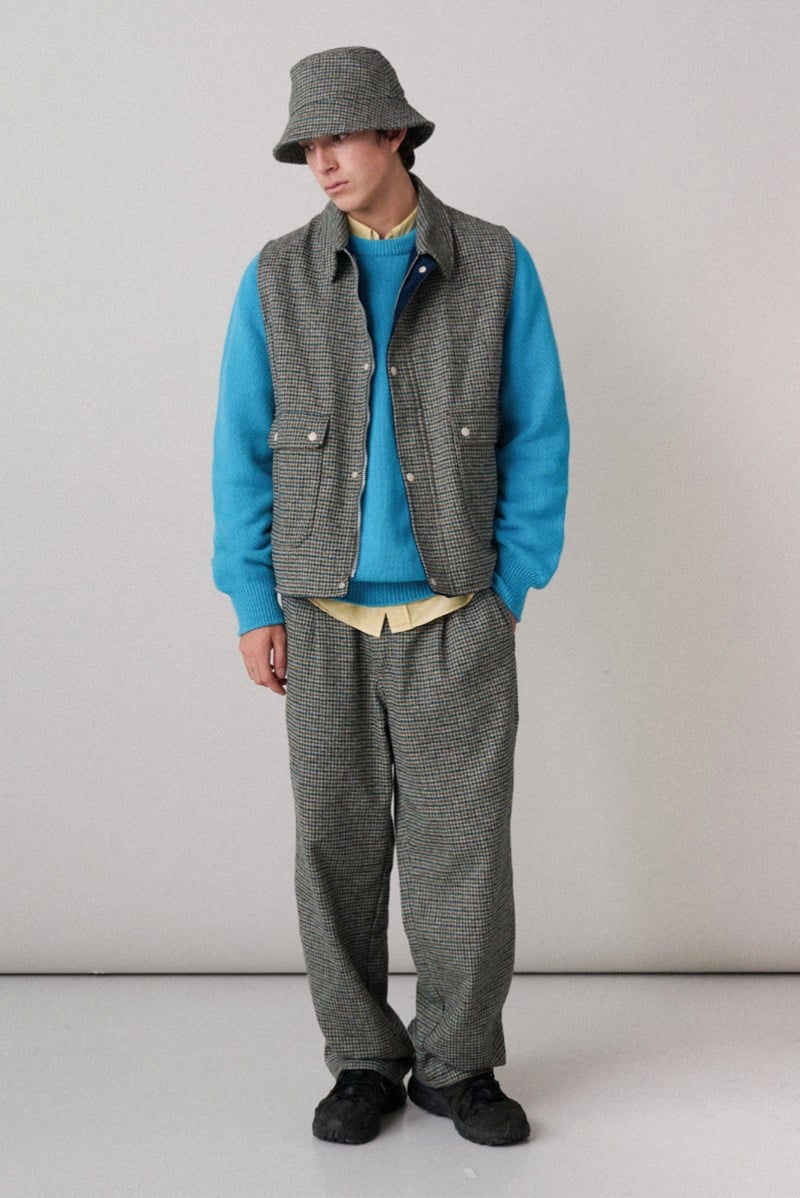 A model in a checked bucket hat and teal sweater layered under a matching checked vest and trousers, for Howlin's fall-winter 2026 collection