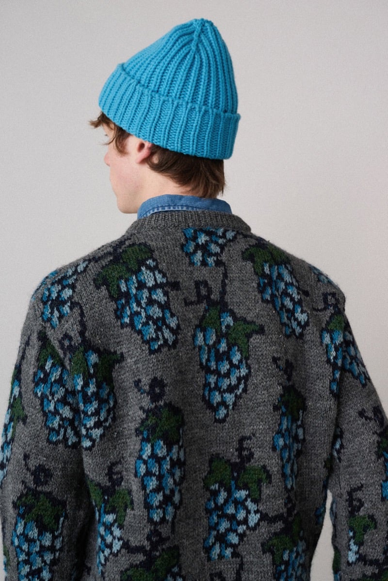 A back view of a model in a gray sweater knitted with a grape motif, styled with a ribbed beanie, for Howlin's fall-winter 2026 collection