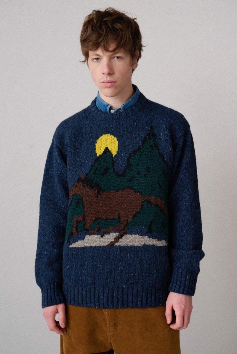 A model in a blue sweater knitted with a horse-and-mountain motif under a moon, for Howlin's fall-winter 2026 collection