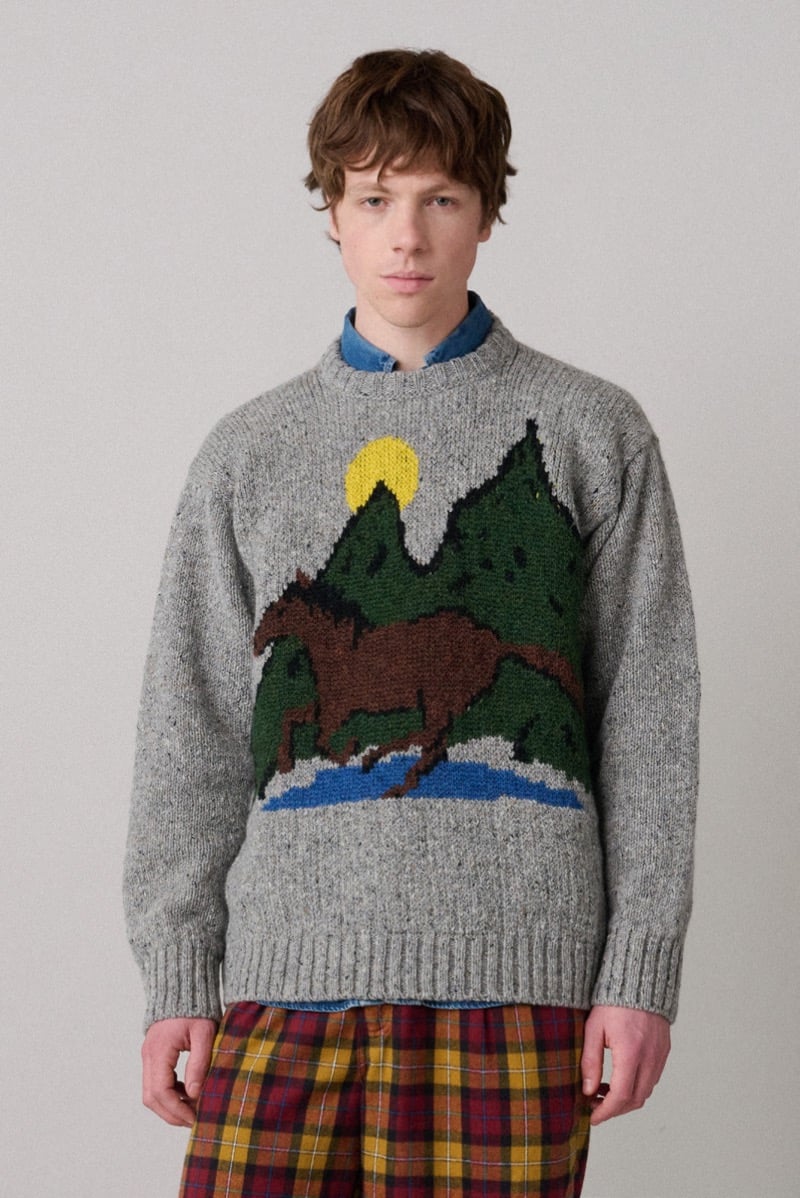 A model in a gray sweater knitted with a horse-and-mountain motif, paired with plaid trousers, for Howlin's fall-winter 2026 collection