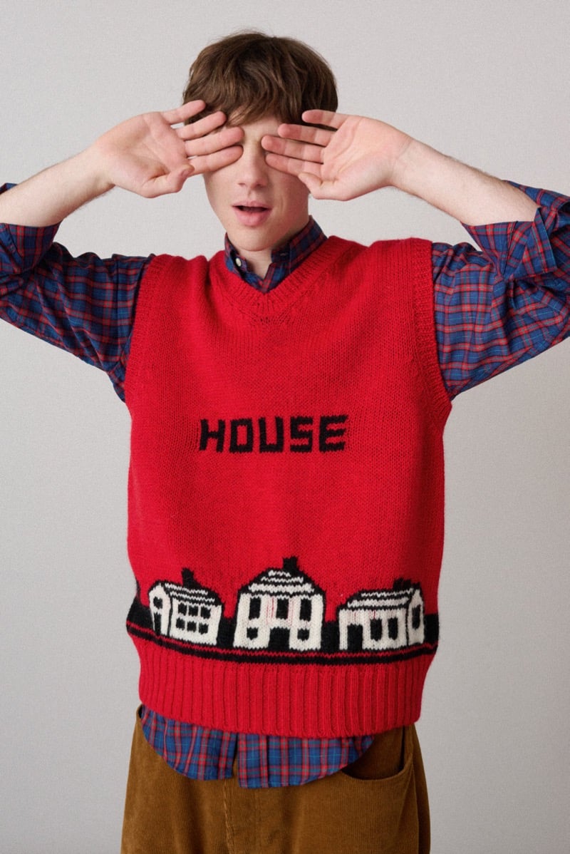 A model in a red sweater vest with a knitted 'House' motif, worn over a plaid shirt, for Howlin's fall-winter 2026 collection