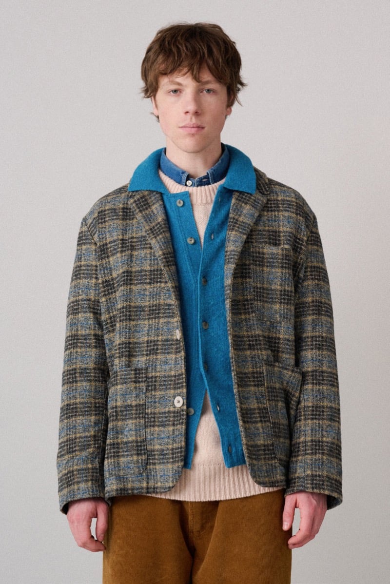 A model in a plaid wool jacket layered over a teal cardigan and a lighter knit sweater, for Howlin's fall-winter 2026 collection