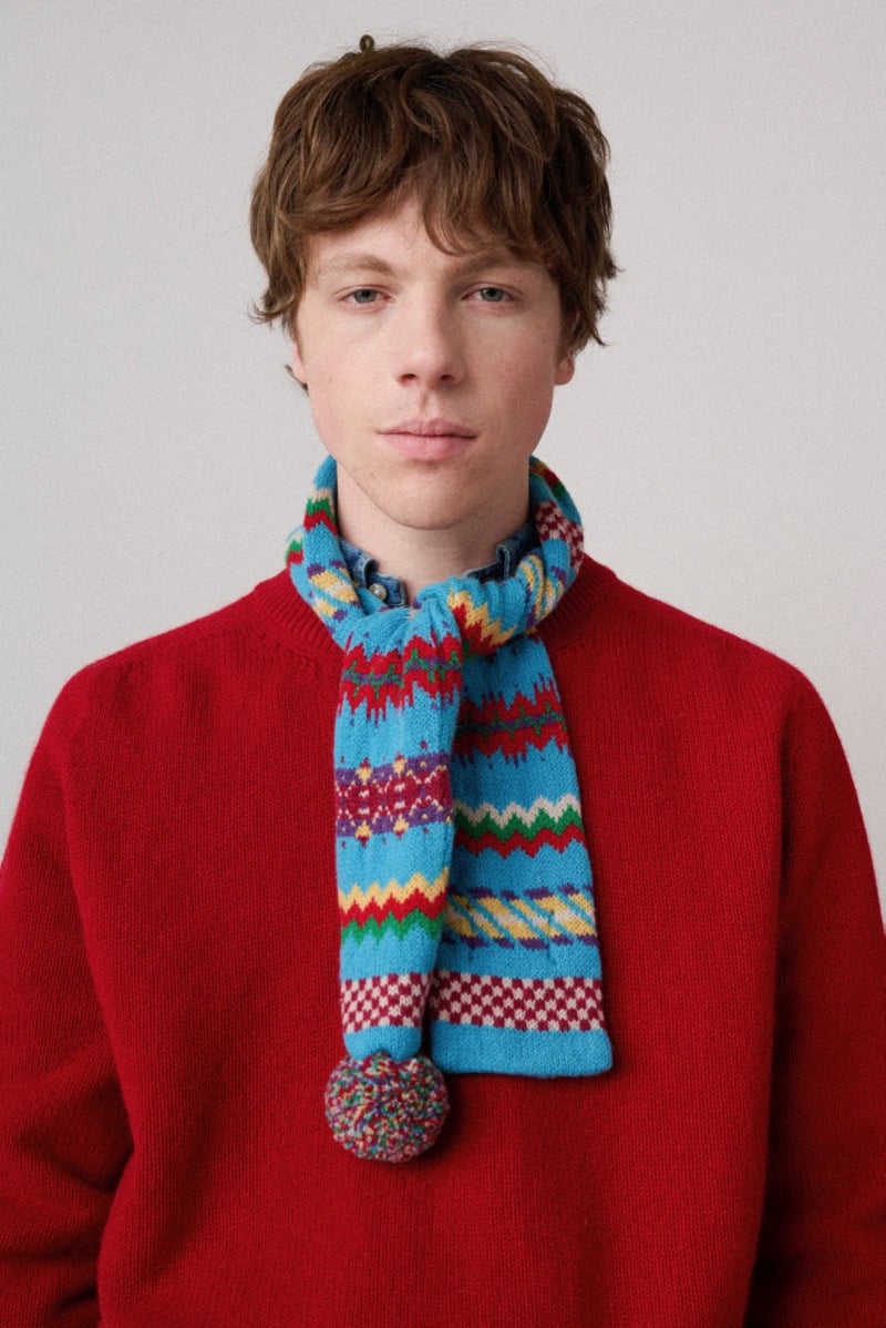 A model in a red sweater with a colorful patterned scarf and pom-pom tied around the neck, for Howlin's fall-winter 2026 collection