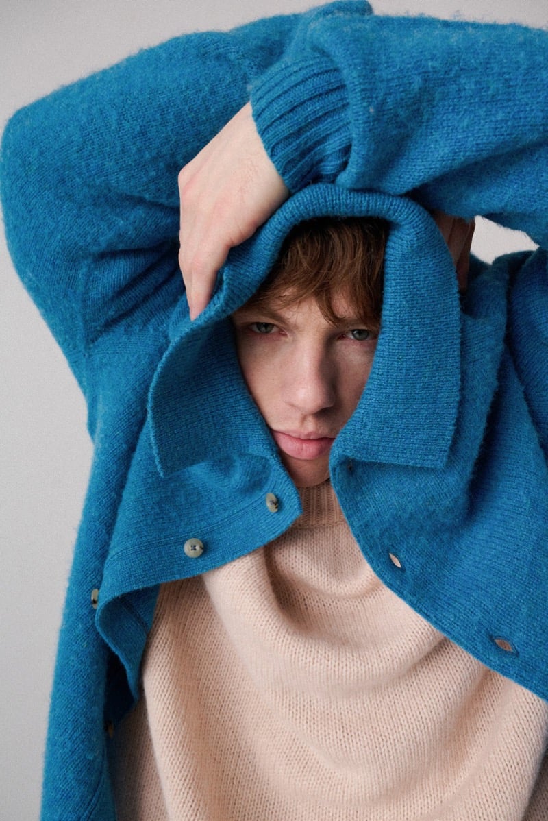A close-up of a model pulling a teal sweater up over his head, with a lighter knit sweater underneath, for Howlin's fall-winter 2026 collection