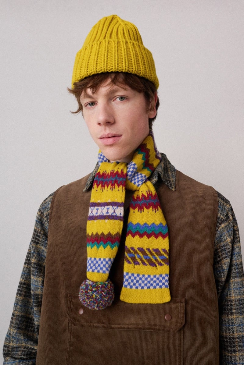 A model in a yellow beanie and a colorful patterned scarf with a pom-pom, layered over a corduroy vest, for Howlin's fall-winter 2026 collection