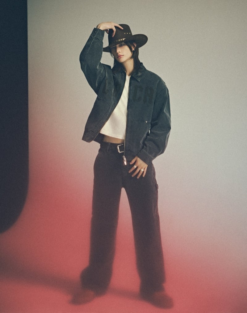 Hyunjin poses in studio with a red glow behind him, wearing a dark GUESS canvas jacket over a cropped pale tee with dark trousers and a brown cowboy hat, for GUESS fall 2026