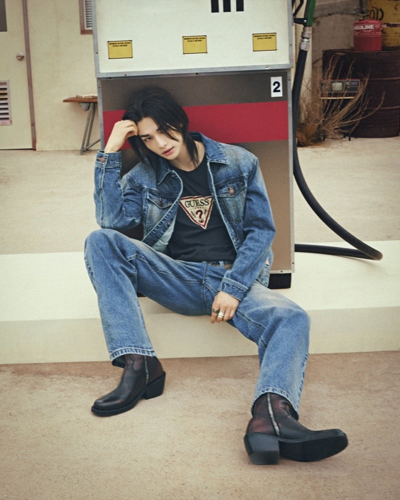 Hyunjin sits against a vintage gas pump in a denim jacket and a dark GUESS triangle-logo tee with light-wash jeans and dark zip ankle boots, for GUESS fall 2026