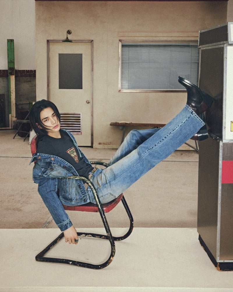 Hyunjin reclines on a tilting cantilever chair with his booted feet propped on a metal machine, wearing a denim jacket over a dark GUESS-logo T-shirt with light-wash jeans, for GUESS fall 2026