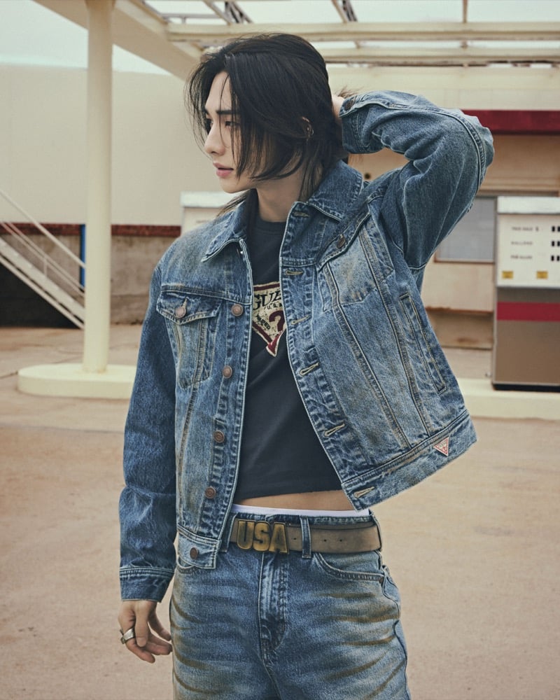 Hyunjin stands at a gas station in a light-wash denim trucker jacket over a dark GUESS triangle-logo tee with light jeans and a USA belt buckle, for GUESS fall 2026