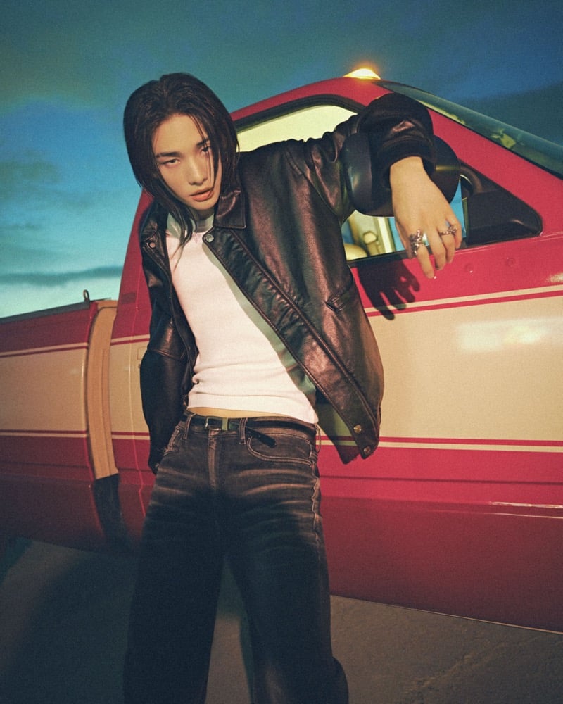 Hyunjin leans against the open door of a red and cream car at dusk, a black leather jacket sliding off one shoulder over a ribbed tank with dark jeans and silver rings, for GUESS fall 2026