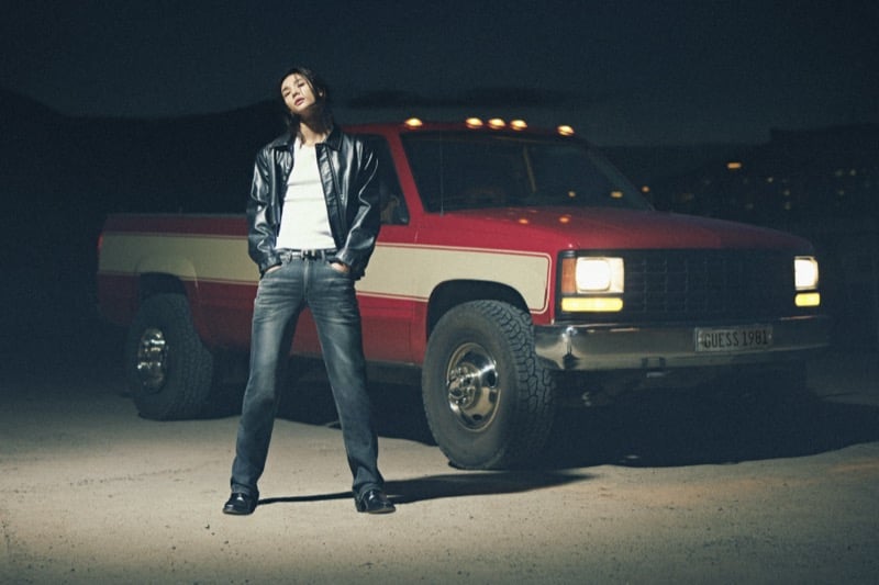 Hyunjin stands with his legs apart in front of a red and cream vintage pickup truck at night, wearing a black leather jacket over a pale ribbed tank with faded jeans and black boots, for GUESS fall 2026
