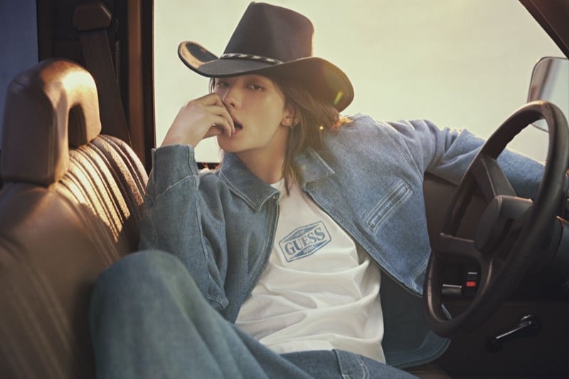 Hyunjin sits in a truck cab with one hand to his mouth, wearing a denim jacket over a white GUESS JEANS T-shirt and a black cowboy hat, for GUESS fall 2026