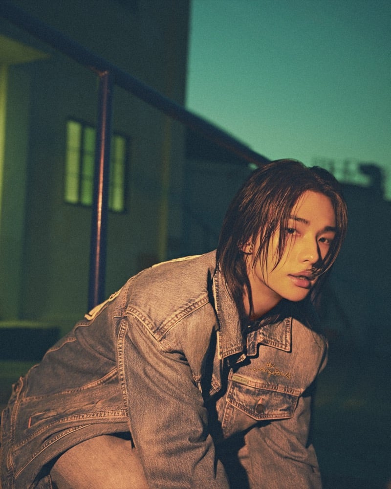 Hyunjin crouches outdoors at dusk in a light-wash denim jacket embroidered Rolling Stones across the chest pocket, for GUESS JEANS x The Rolling Stones