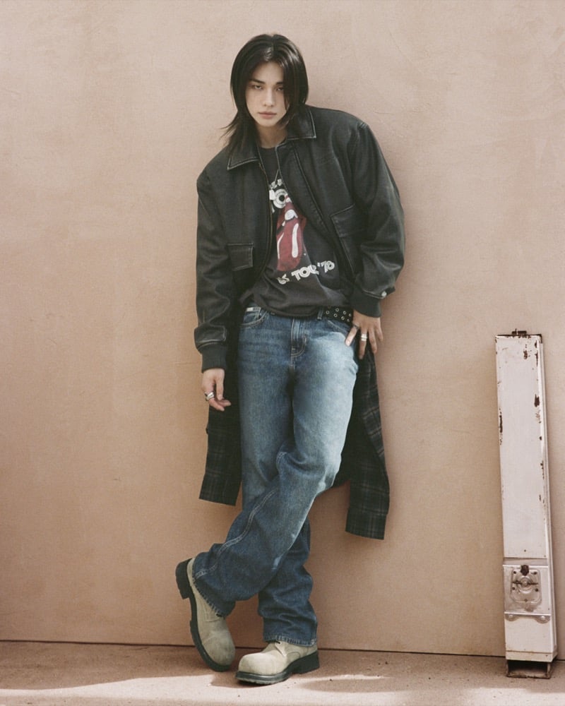 Hyunjin leans against an adobe wall in a black leather bomber over a Rolling Stones T-shirt with baggy light-wash jeans and beige suede boots, for GUESS JEANS x The Rolling Stones