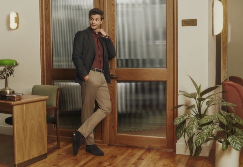 Jack Quaid in a charcoal blazer, burgundy button-up, and tan chinos leans against a glass-paned door marked 'Weekday Warrior' for Bonobos' fall 2026 campaign