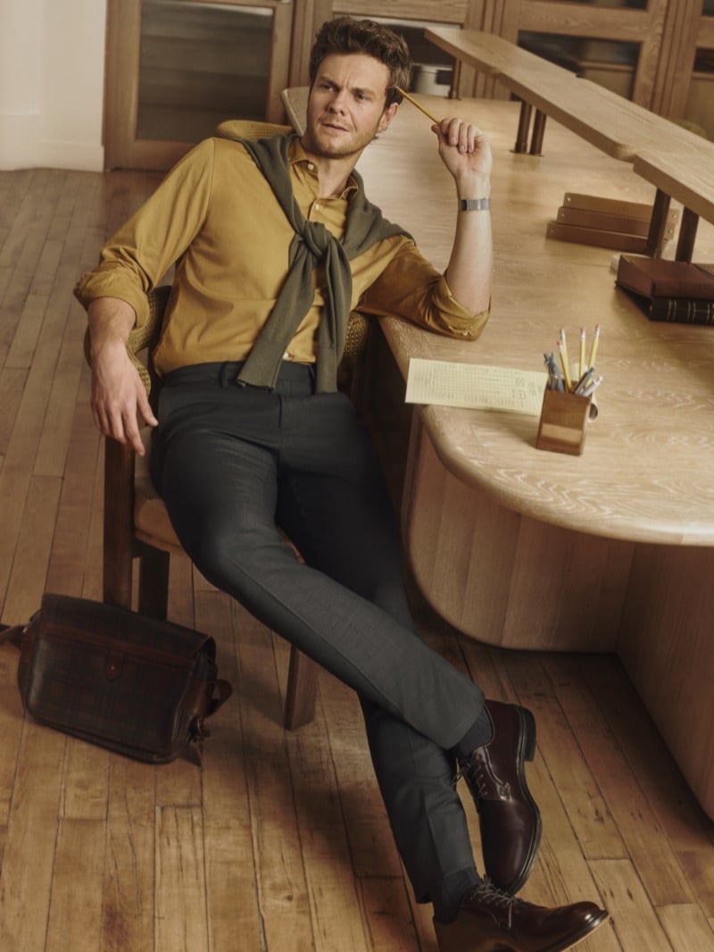 Jack Quaid twirls a pencil at a desk in a mustard button-up with an olive sweater tied over his shoulders, for Bonobos' Weekday Warrior campaign