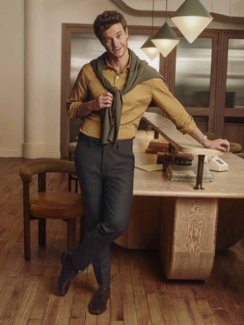 Jack Quaid stands by a desk in a mustard button-up with an olive sweater tied over his shoulders and charcoal trousers, for Bonobos' Weekday Warrior campaign