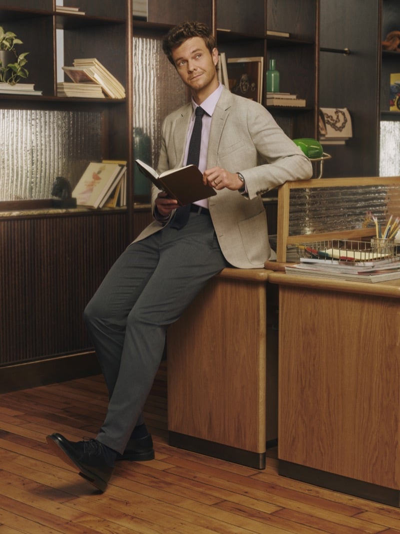 Jack Quaid sits on a desk reading a book, wearing a flecked oatmeal blazer, pale pink shirt, and tie with charcoal trousers, for Bonobos' Weekday Warrior campaign