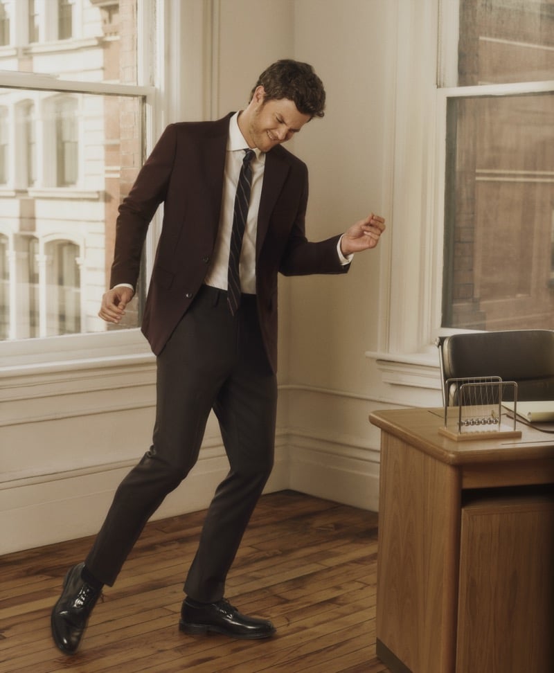 Jack Quaid dances beside an office window in an oxblood blazer, white shirt, and striped tie, for Bonobos' Weekday Warrior campaign