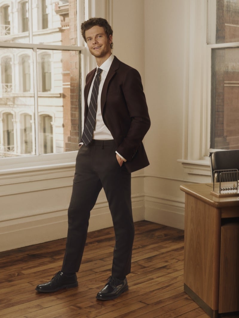Jack Quaid smiles by a window in an oxblood blazer, white shirt, and striped tie, for Bonobos' Weekday Warrior campaign