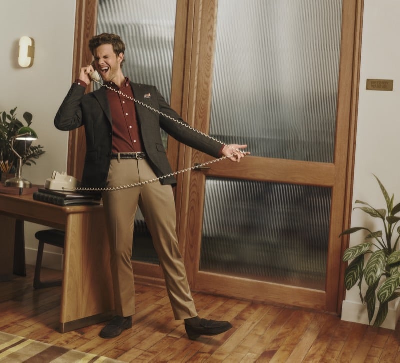 Jack Quaid talks into a corded phone in a charcoal blazer, burgundy button-up, and tan chinos, for Bonobos' Weekday Warrior campaign