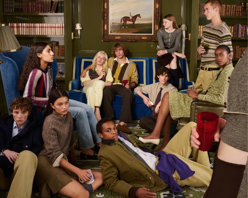 A crowded house party gathers around a library sofa, models layered in rollneck sweaters, rugby stripes, and an olive barn jacket, for J.Crew's Eckhaus Latta collection