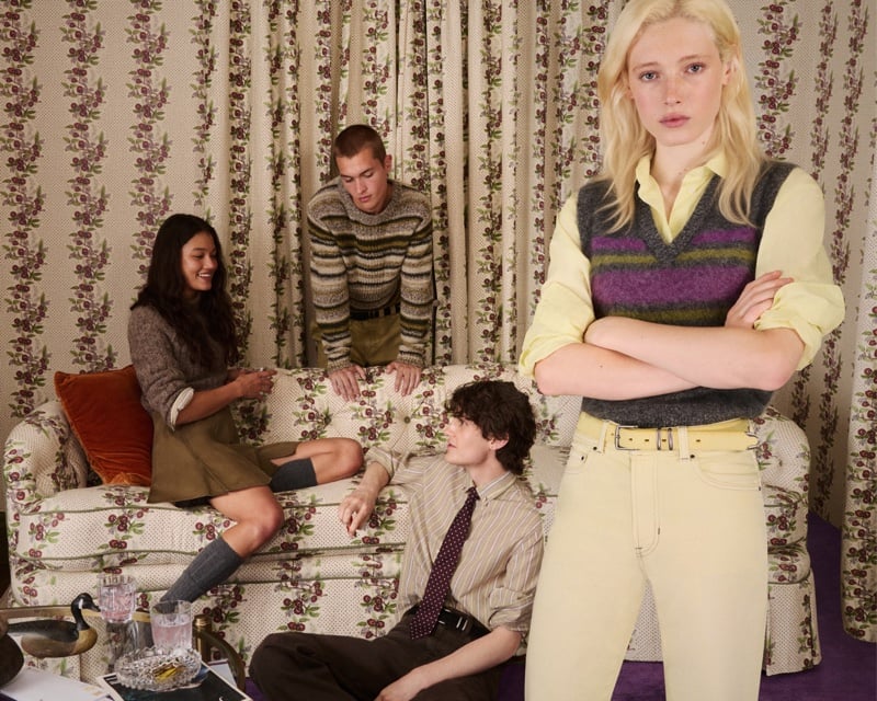A model crosses her arms in a striped mohair sweater vest over a yellow button-up shirt, standing before a floral-curtained room where two models sit on a patterned sofa, for J.Crew's Eckhaus Latta collection