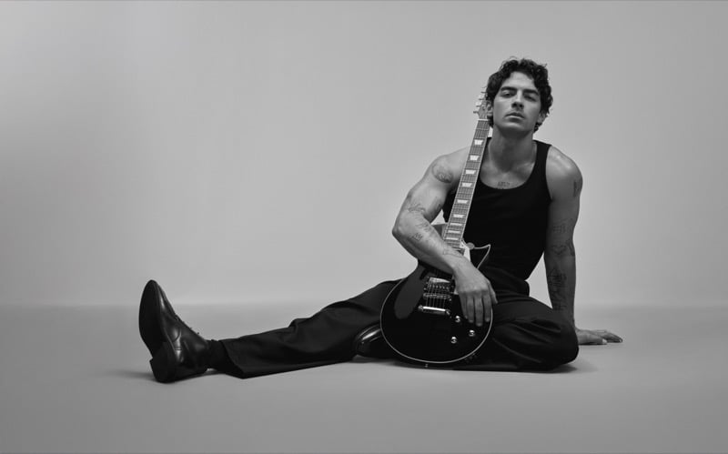 Joe Jonas sits on the floor of a studio with an electric guitar resting against his shoulder, wearing a tank top, trousers, and leather lace-ups for Kenneth Cole INSPIRE.