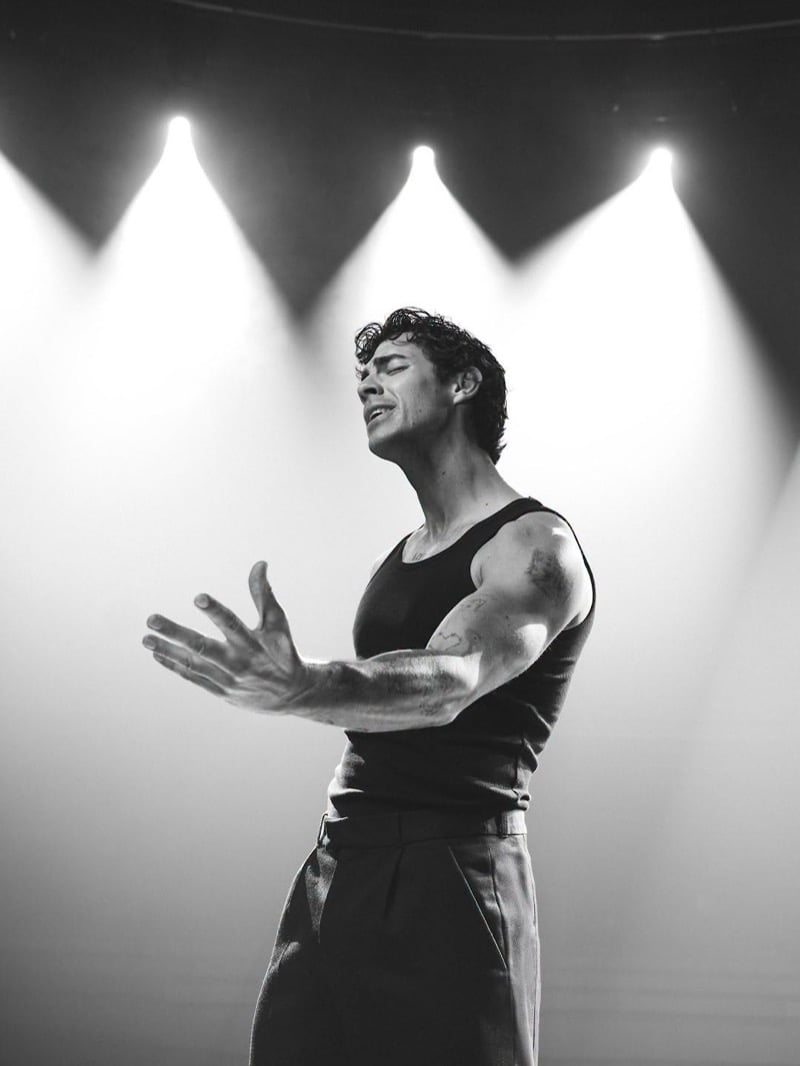 Joe Jonas sings with his eyes closed and one arm outstretched beneath stage lights in a tank top and pleated trousers for the Kenneth Cole INSPIRE campaign.