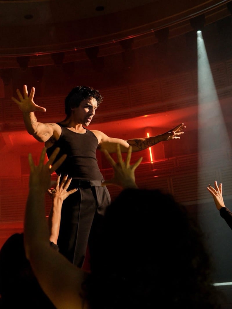 Joe Jonas stretches his arms out on stage in a tank top and belted trousers as fans reach toward him under red light for the Kenneth Cole INSPIRE campaign.