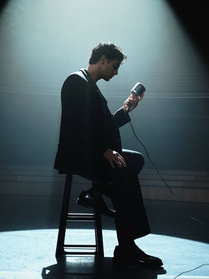 Joe Jonas sits on a wooden stool under a stage spotlight in a suit, looking down at a microphone, for the Kenneth Cole INSPIRE campaign.