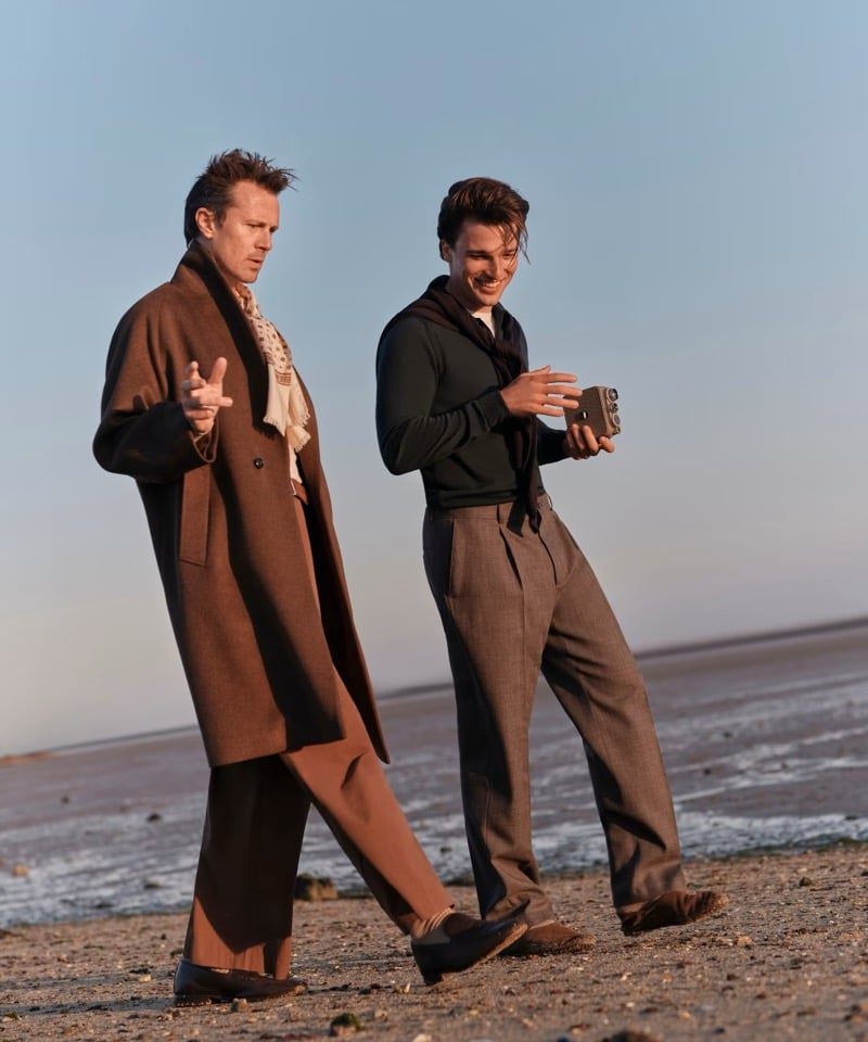 Robert Montgomery and Kit Clarke walk along a beach at dusk for John Smedley, Montgomery in a long brown overcoat over a patterned scarf and wide trousers, Clarke in a dark green zip-through knit with a scarf draped over the shoulders and grey pleated trousers, holding a small vintage film camera