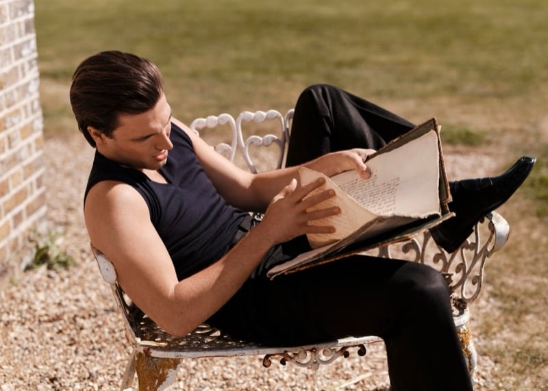 Kit Clarke reclines on a white wrought-iron garden bench reading a worn hardcover book, wearing a dark sleeveless knit and dark tailored trousers with black loafers for John Smedley
